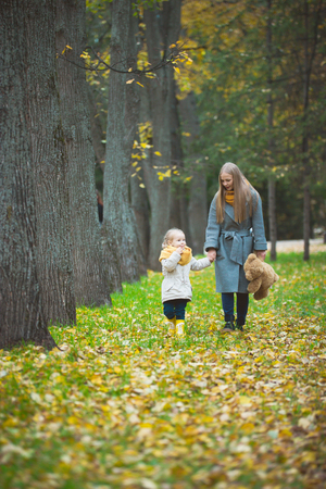 Mother and little daughter blonde with Teddy bear walking in autumn park, yellow lives around, telephotoの写真素材