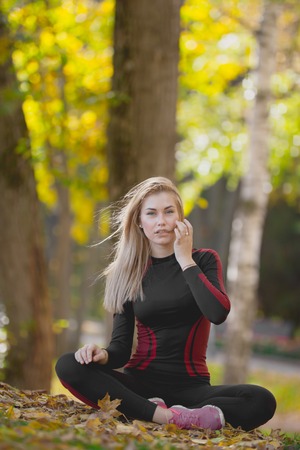 Young attractive fitness female sitting cross-legged at workout in autumn park, straightens her waving in the wind hair, telephotoの写真素材