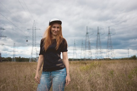 Young energy happy woman standing at the camera on yellow field with power pylons, cloudy autumn