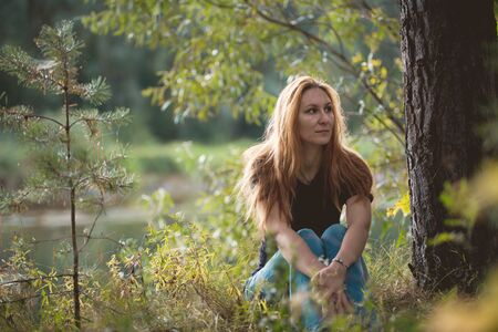 Beautiful red-haired girl sitting near river at forest, telephotoの写真素材