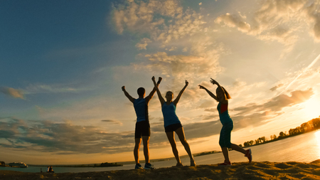 Group of young people - athletes - two girls and a guy are have triumph on the mountain, near river at dusk, silhouetteの写真素材