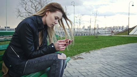 Dissapointed girl with long blonde hair in leather jacket straightens hair use gadget sitting on the bench in the wind, telephotoの写真素材
