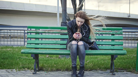 Dissapointed girl with long blonde hair in leather jacket straightens hair use gadget sitting on the bench in the wind listen headphones, wide angle, front view, telephotoの写真素材