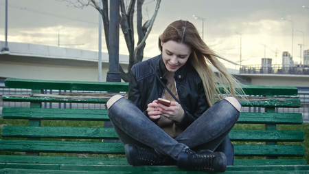 Cute happy girl with long blonde hair in leather jacket straightens hair use gadget sitting on the bench in the wind, close upの写真素材
