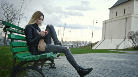 Cute happy girl with long blonde hair in leather jacket straightens hair use gadget sitting on the bench, horizontalの写真素材