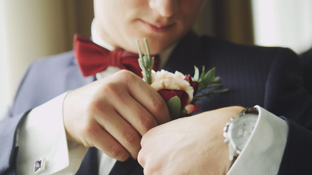 Fashion young man in bow tie and clock - groom's hand arranging boutonniere flower on suit, close upの写真素材
