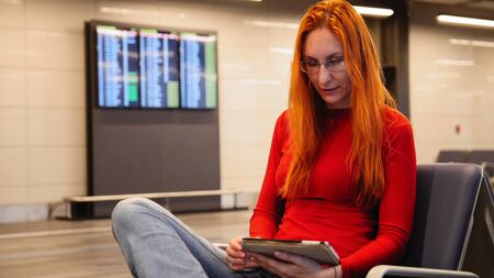 Young attractive woman with red hair and glasses use gadget in airport departure lounge, close upの写真素材