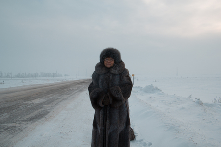 Portrait of senior woman fur coat and hat standing in cold winter snow covered road, telephotoの写真素材