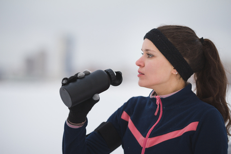 Sport cute woman drinks water during jogging outside at snow winter dayの写真素材