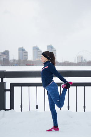 Young female runner doing flexibility exercise for legs before or after running at snow winter promenade, verticalの写真素材