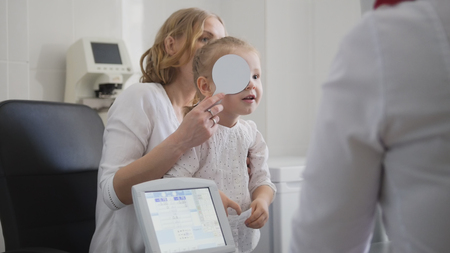 Blonde little girl closes one eye - Optometrist in clinic checking little childs visionの写真素材