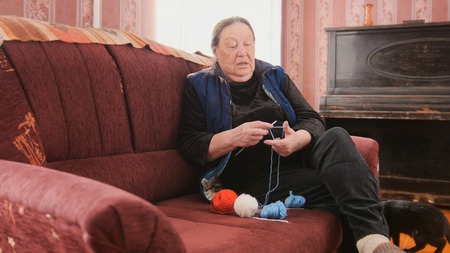 Senior lady sitting on the sofa and knits colourful wool - pensioners hobby at homeの写真素材