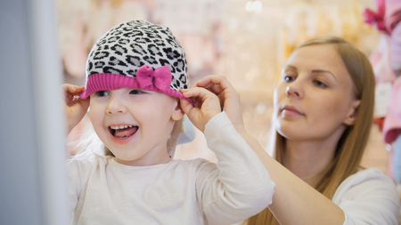 Little happy girl with mommy doing shopping and buying pink hat in kids dress storeの写真素材