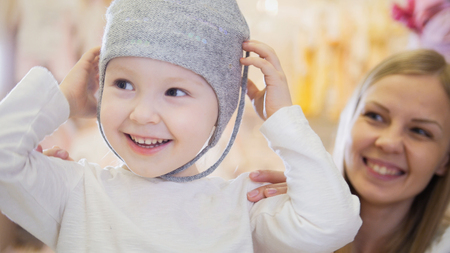 Little blonde baby girl with mother doing shopping and buying hat in kids dress storeの写真素材