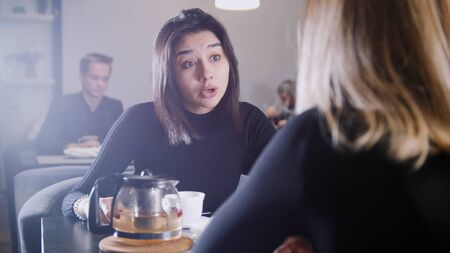 Black haired young woman with black hair drinking coffee and talking with girlfriend in the cafeの写真素材