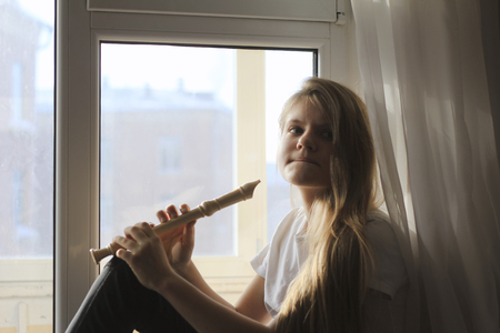 Young cute teen girl playing on flute sitting on windowsill at homeの写真素材