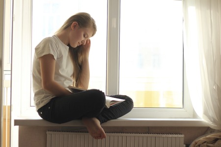 Young cute blonde teen girl sitting on windowsill and reading a bookの写真素材