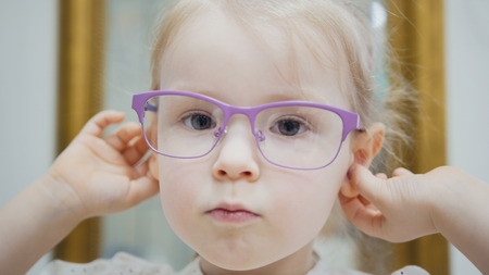Little girl tries fashion medical glasses near mirror - shopping in ophthalmology clinicの写真素材