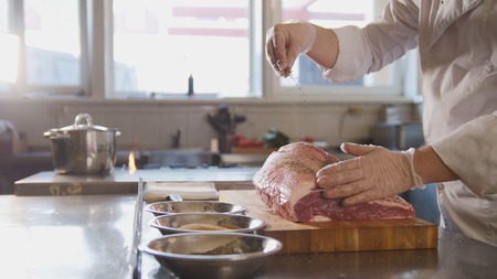 Butcher adding spices into large piece of fresh raw meat lying on a wooden board in a commercial kitchenの写真素材