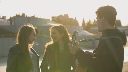 Two girls smiling while guy playing on the guitar at the sunset outdoors