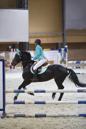 Slim young woman on the black horse galloping at show jumping competitionの写真素材