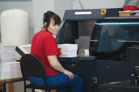 Adult female worker with a stacks of paper in front of print press equipmentの写真素材
