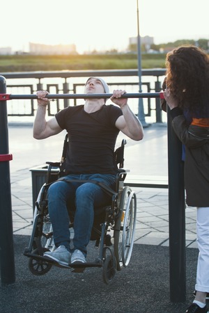 Strong disabled man in wheelchair pulled up on the bar next to young woman at sunsetの写真素材