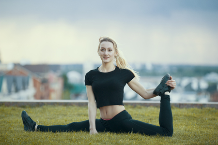 Happy young woman on the grass performs twine with the bent leg, on background cityscapeの写真素材