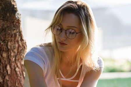 Portrait of young attractive woman in eyeglasses near the tree in park at sunny dayの写真素材