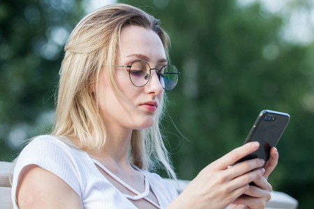 Portrait of young attractive woman in eyeglasses uses a smartphone outdoors in summer dayの写真素材