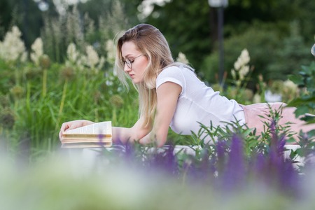 Young beautiful woman reading a book lying on the bench around the grass and flowers at summer dayの写真素材