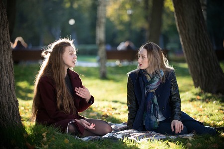 A beautiful long-haired girl tells a story to her friend. They sit on a plaid in the autumn parkの写真素材