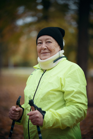 An elderly woman in a green down jacket is standing in a park with sticks for nordic walkingの写真素材