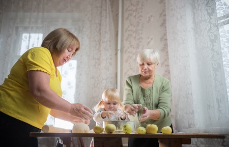 Two elderly woman making apple little pies at home. Sifting flour with little blonde girlの写真素材
