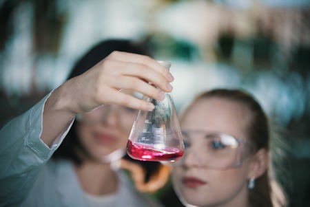 Chemical laboratory. Two young woman holding a flask with pink liquid in it. Flask in focusの写真素材