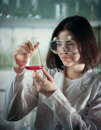 Chemical laboratory. Young woman in work glasses looking at the flask