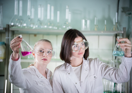 Chemical laboratory. Two young pretty woman holding different flasks with liquids in it. Looking at...