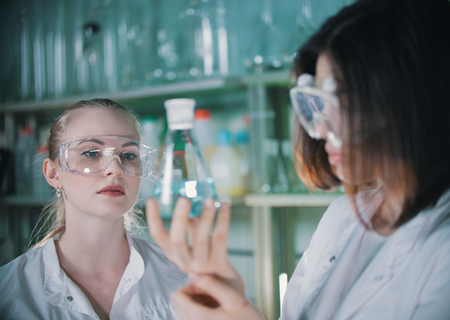 Two young woman in chemical lab holding a flasks with clear liquid in it. Blonde woman in focusの写真素材