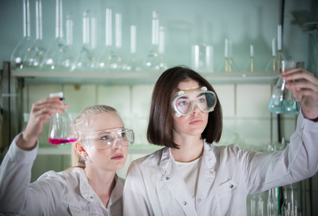 Chemical laboratory. Two young woman holding different flasks with liquids in it