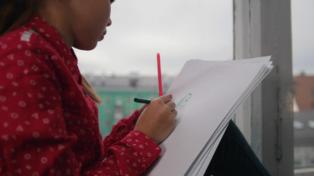 A little girl sitting on the window sill and drawing, smiling on the backround of the city. Focus on paperの写真素材