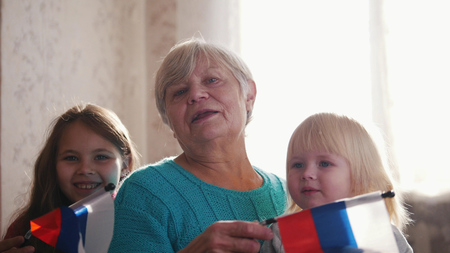 A nice girls and their grandmother enjoy sunny morning. Good time at home. Flag of Russia in hands...