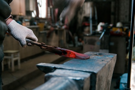 Blacksmith working with red hot metal workpiece of new axe on the anvil at the forge. Focus on handsの写真素材