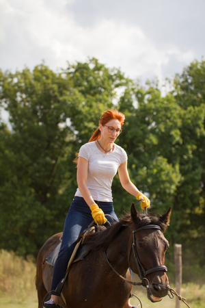 A redhead woman in white t-shirt riding a horse in nature. Mid shotの写真素材
