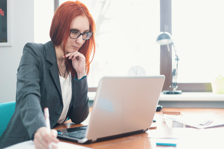 A ginger woman in glasses sitting at office and looking at the laptopの写真素材