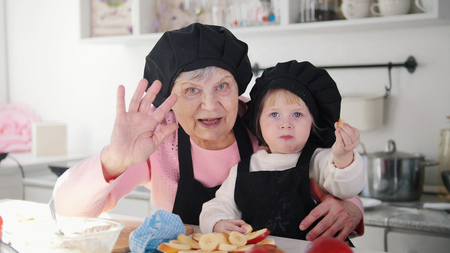 An old woman with a little girl waving hands and looking in the camera in the kitchen.の写真素材