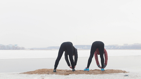 Two slim women standing on the snowy beach and warming up for the running. Bending down