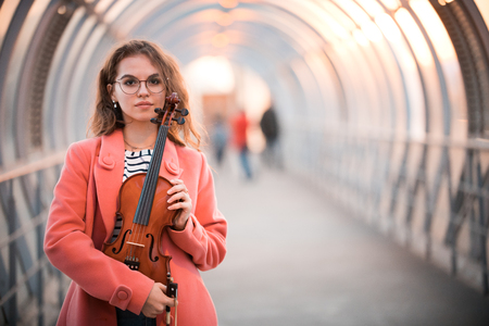 Young woman in glasses standing on the upper pedestrian crossing in a sunset holding a violinの写真素材