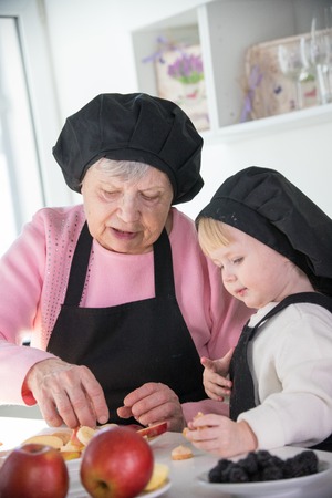 An old woman with a little girl standing in the kitchen preparing fruits for a serving
