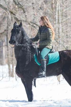A winter forest. A woman with long wearing green jacket hair riding a dark brown horse