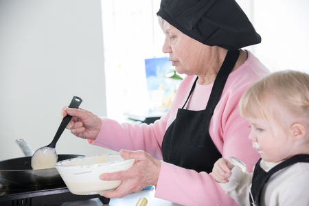 Family making pancakes. A little girl sloppy eating a sour cream while her grandmother put the dough in the panの写真素材